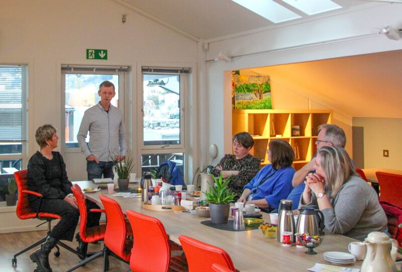 Bilete visar 5 personar som sit rundt eit bord på frokostmøte, medan ein står på enden av bordet og heldt foredrag