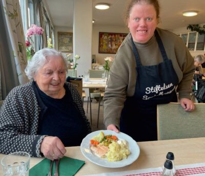 Elisabeth smiler til kamera når ho serverer middag til 100 år gamle Johanna
