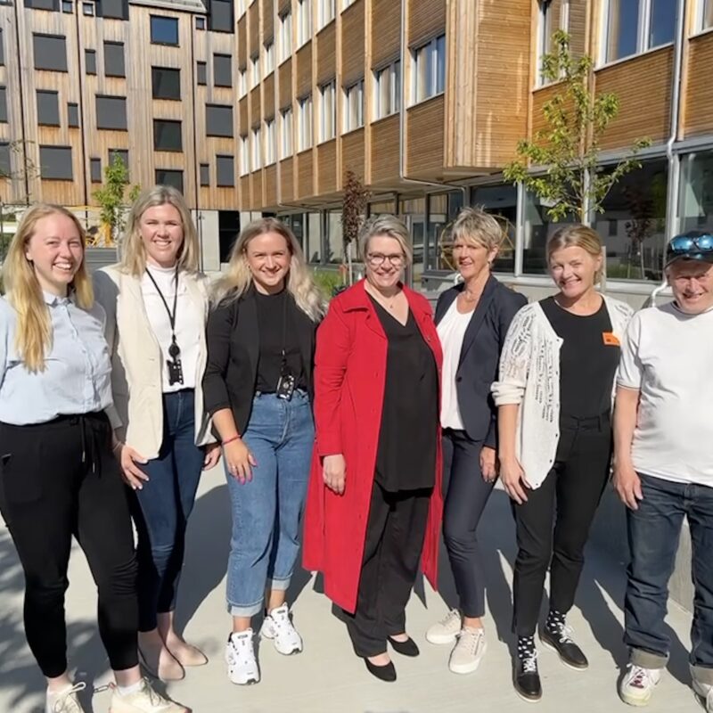 Kveik saman med Arbeids- og inkluderingsminister Marte Mjøs Persen og andre aktørar. Dei smilar og ser i kamera. I bakgrunnen ser me fasaden til Scandic Hotel på Voss.