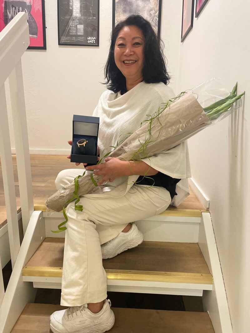 Mari Skjervheim feirar 25 år med Kveik AS, ho sitt i trappa på Kveik og smilar, med blomar og gåve i handa.