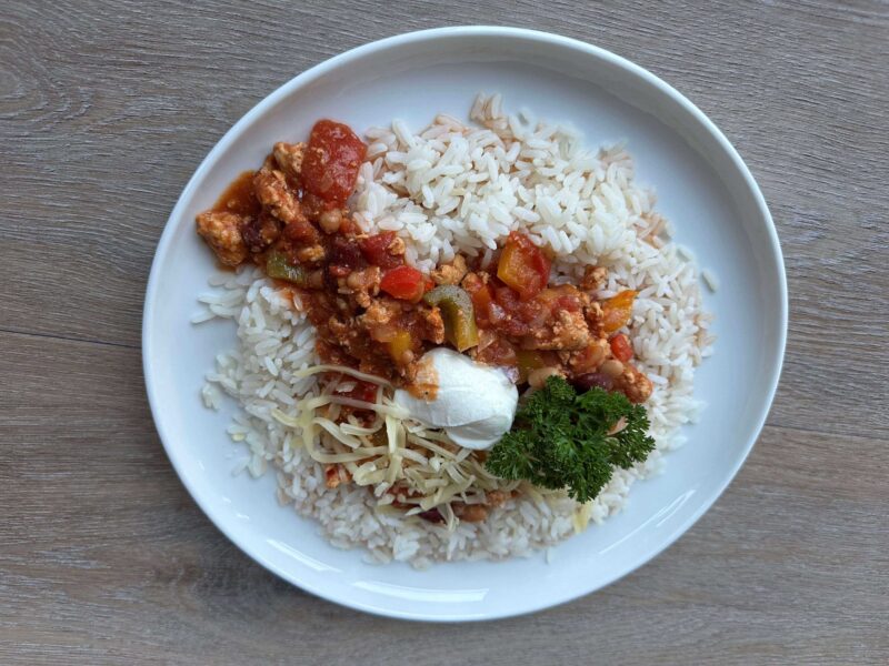 Matretten chili con carne på eit kvitt fat med ris, rømme og ein persilledusk