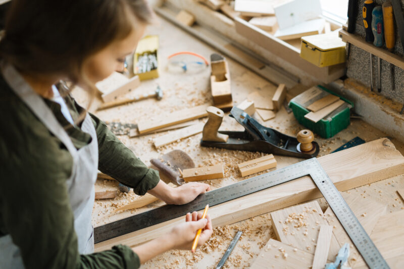 Ei kvinne jobbar med oppmåling av ein planke på snekkerbenken sin. Rundt ho er det spon og mange verktøy.