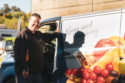 Ein ansatt i Kveik smiler og ser mot kamera mens han lener seg mot jobbfruktbilen til Kveik. På bilen står det "Korga med meining" i løkkeskrift, og bilen er foliert med fruktbilete.