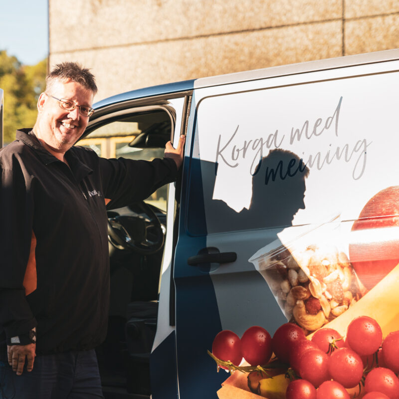 Ein ansatt i Kveik smiler og ser mot kamera mens han lener seg mot jobbfruktbilen til Kveik. På bilen står det "Korga med meining" i løkkeskrift, og bilen er foliert med fruktbilete.