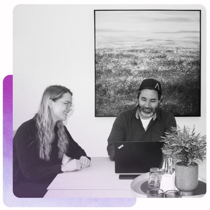 Et bilde av to ansatte i kveik som smiler mot en datamaskin skal vise arbeidsgleden ved arbeidstrening. Bildet er i sort og hvit med en lilla gradering, og bak bildet er en lilla dekorativ kant.